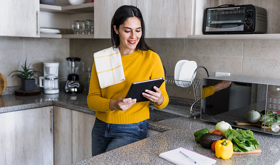 Mujer en la cocina, rodeada de electrodomésticos y mirando su tablet con interés